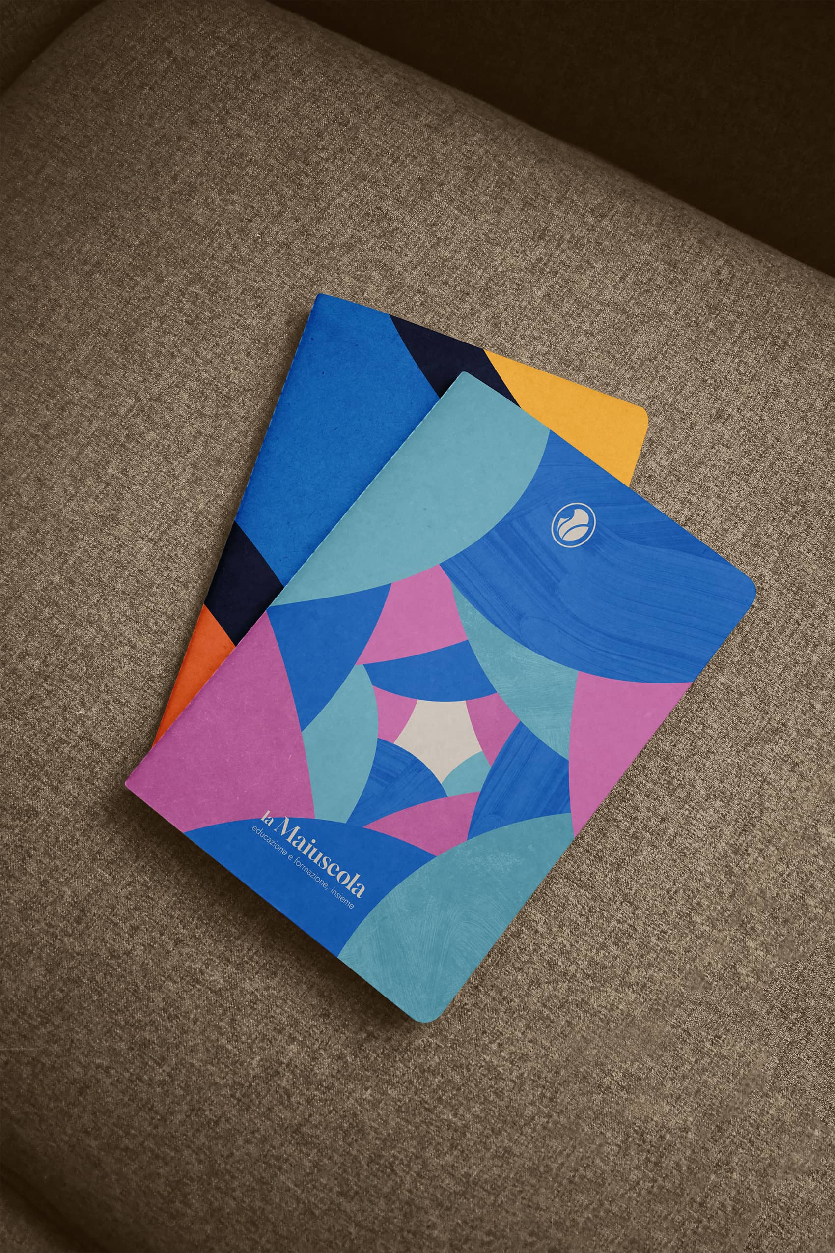 Two colorful notebooks with geometric patterns in blue, pink, orange, and yellow placed on a brown fabric surface.