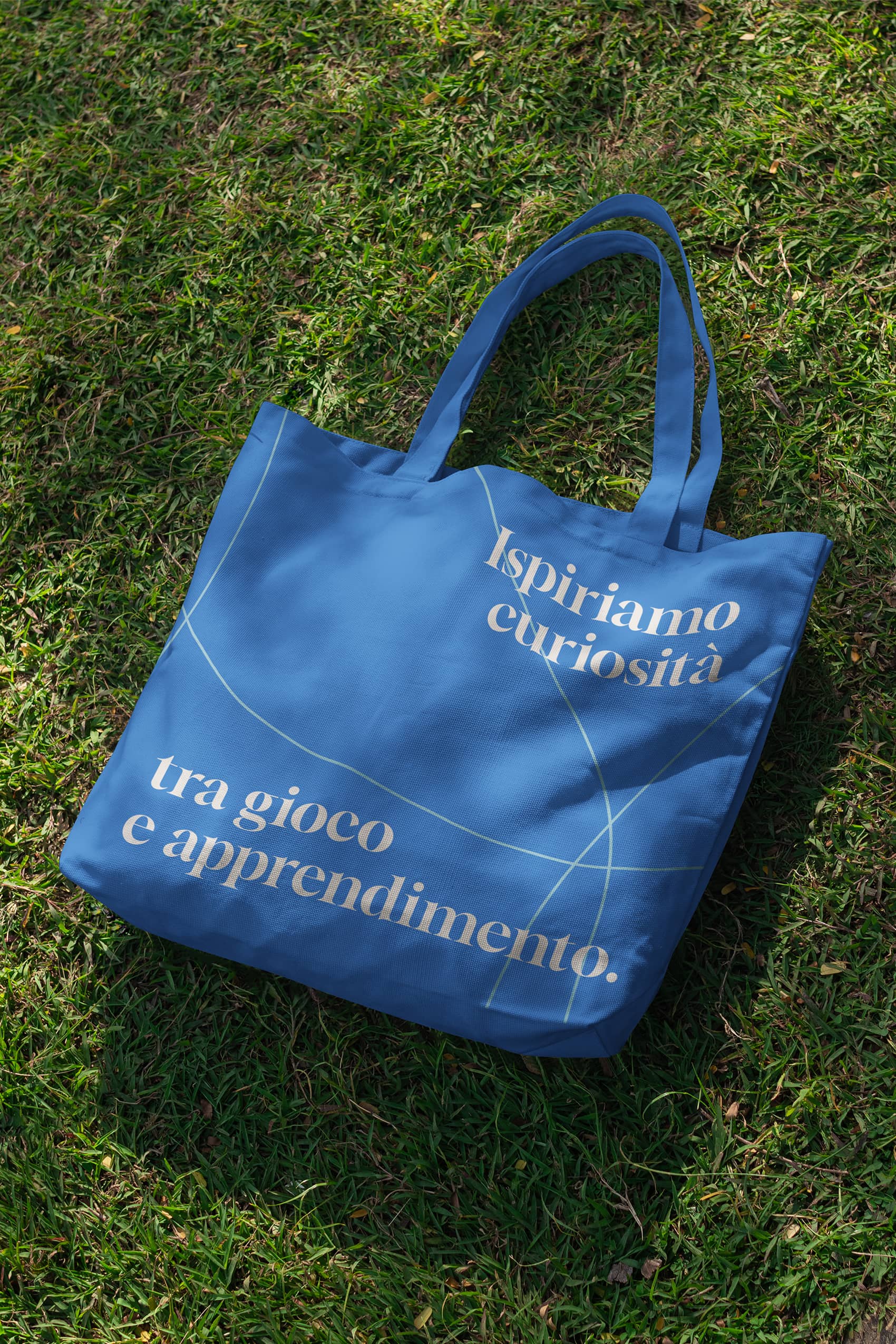 Blue tote bag with Italian text 'Ispiriamo curiosità tra gioco e apprendimento' lying on green grass.