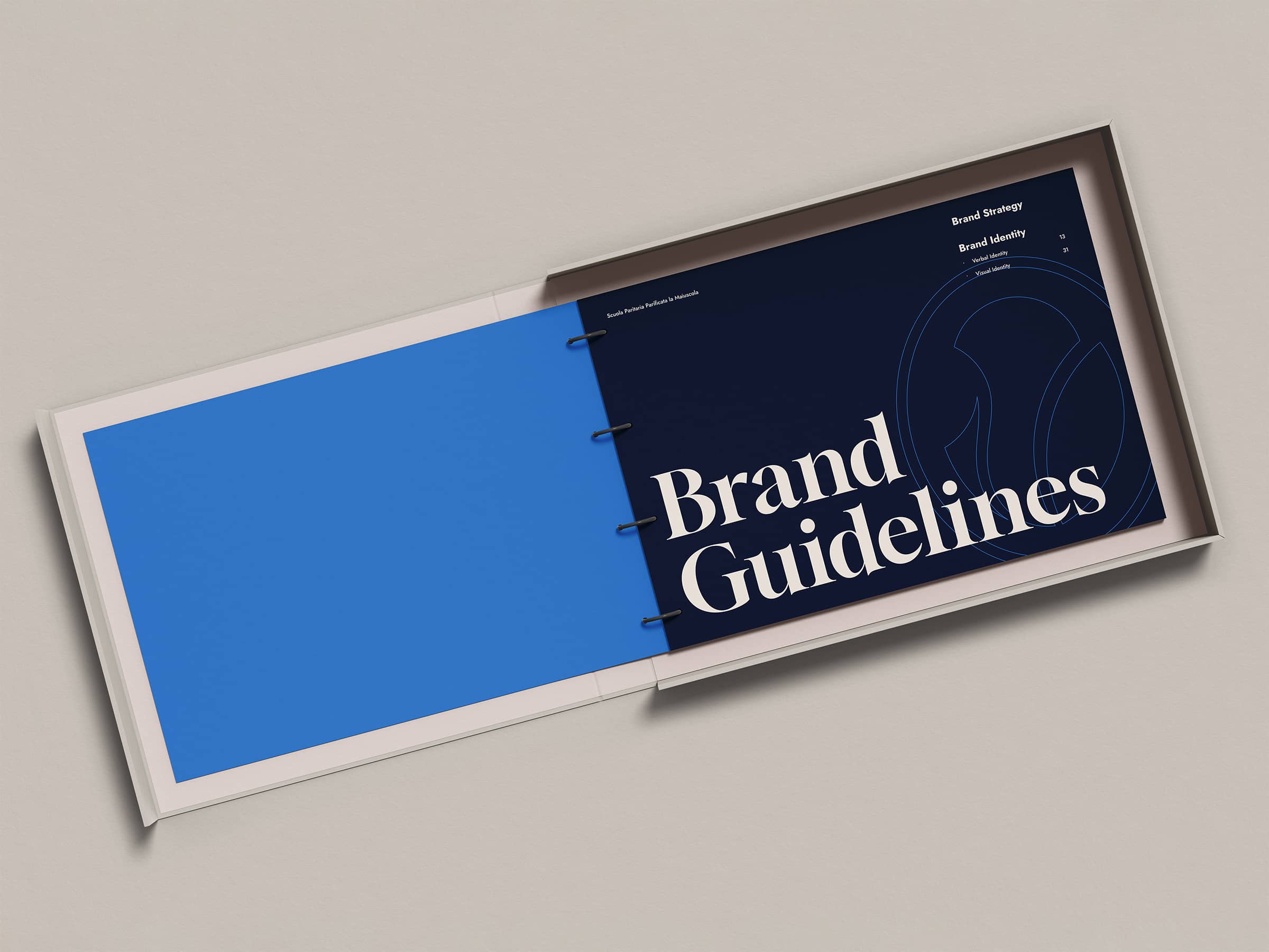 Open booklet on a beige surface showing a blue page and a dark blue page titled 'Brand Guidelines' with sections on Brand Strategy and Brand Identity.