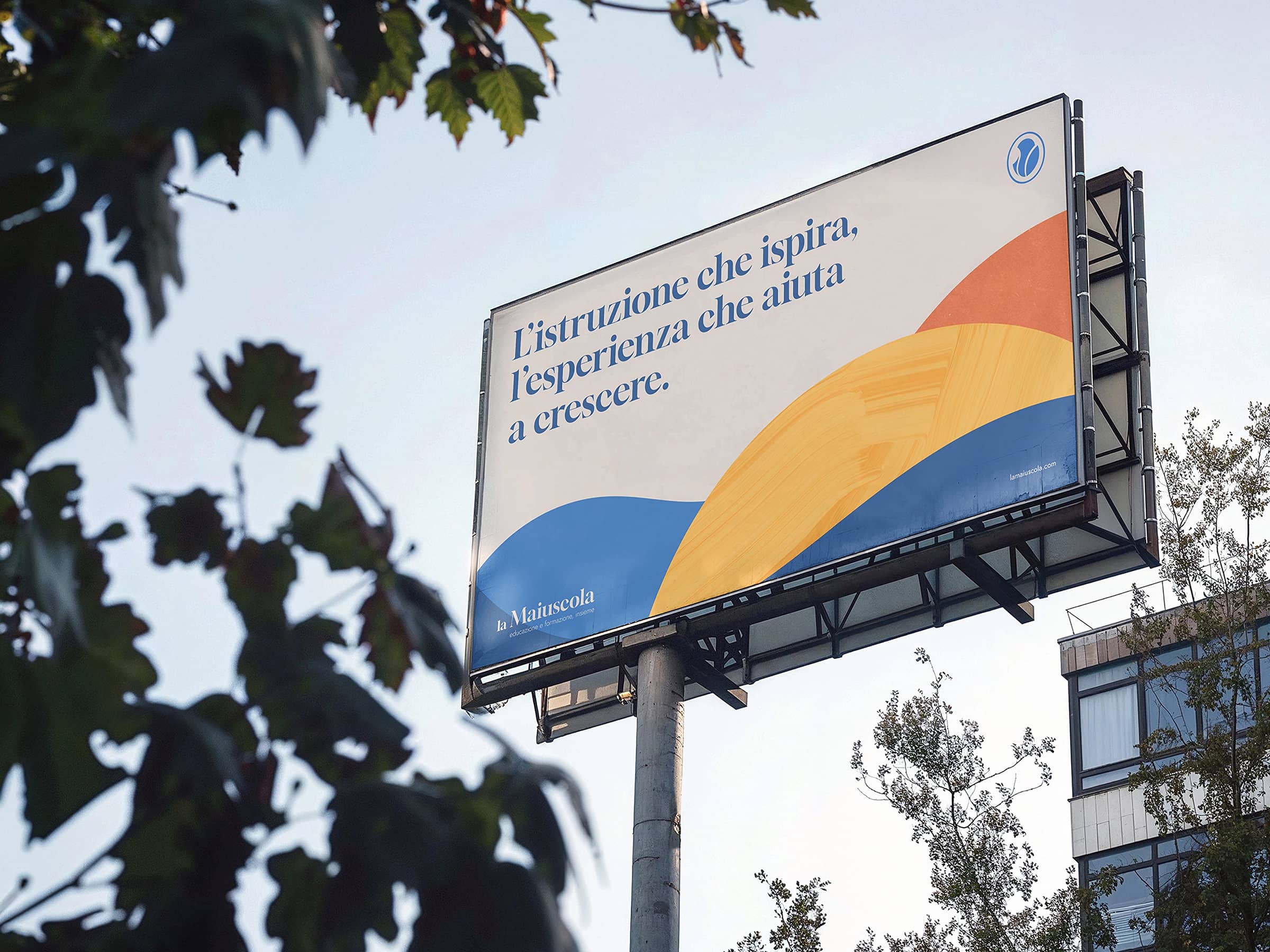 Billboard featuring Italian text about education with abstract blue, yellow, and orange shapes against a pale background, mounted on a tall pole outdoors.