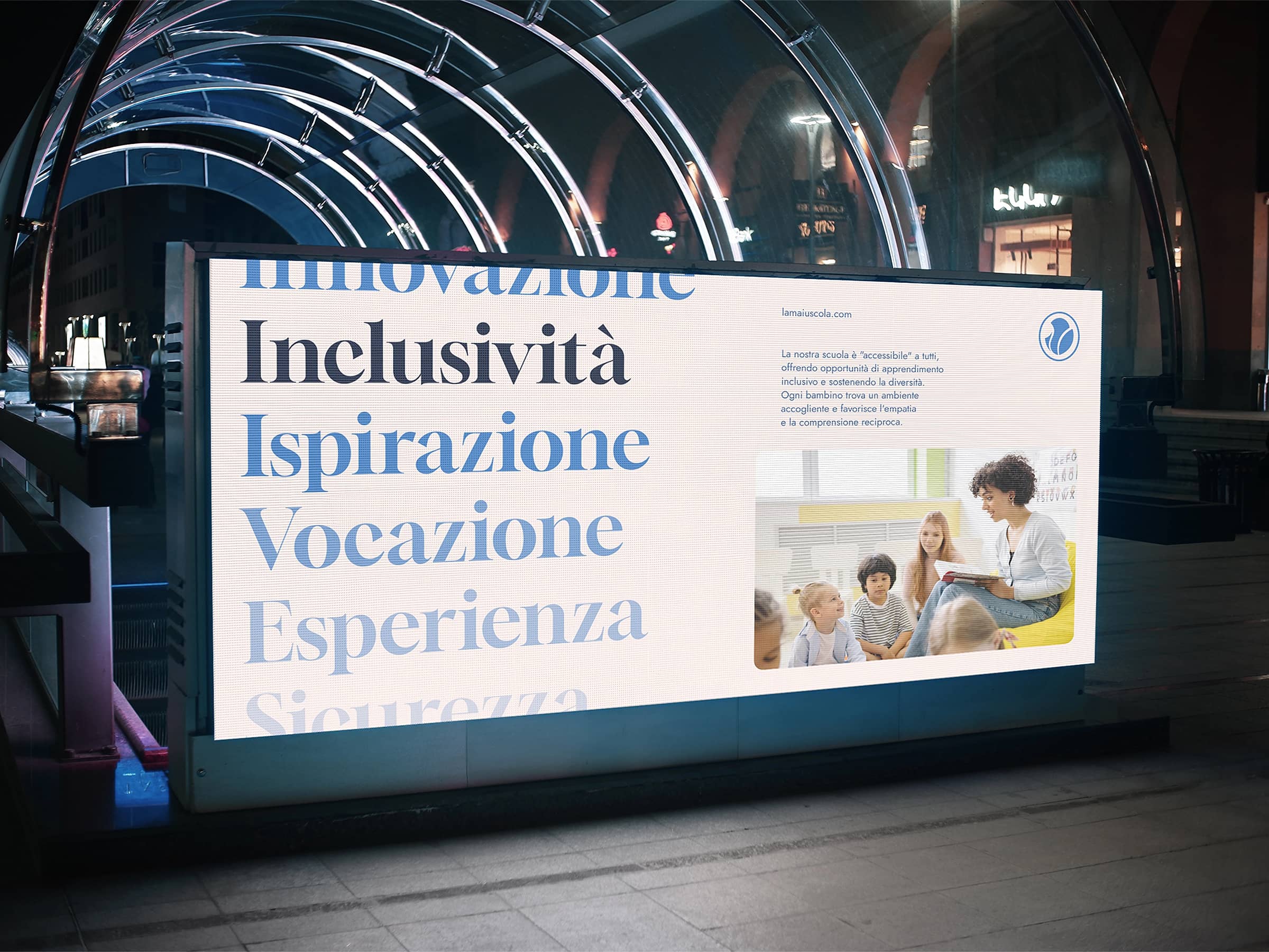 Illuminated billboard in a nighttime urban setting displaying Italian words about inclusivity and a photo of a woman reading to children.
