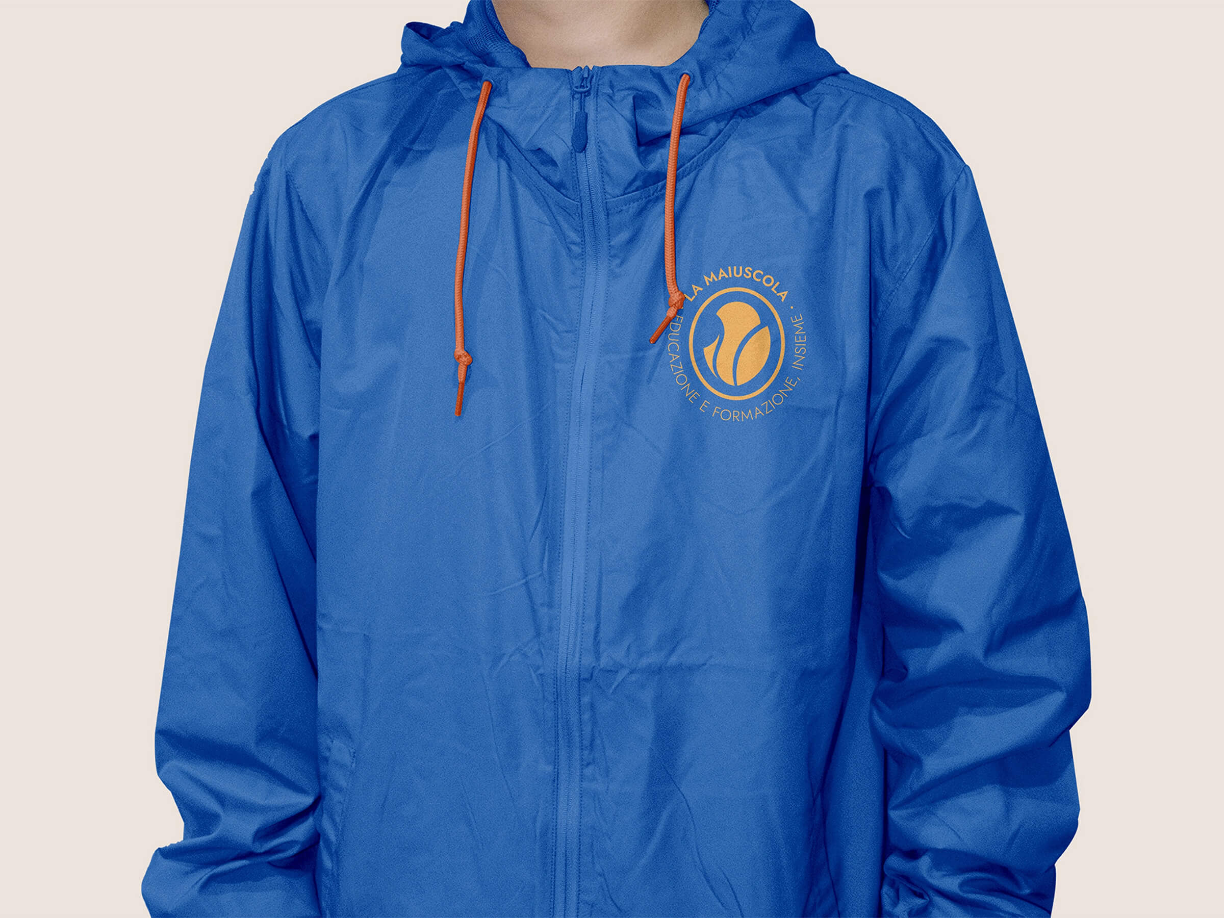 Person wearing a blue zip-up hooded jacket with orange drawstrings and a circular logo on the chest reading 'La Maiuscola Educazione e Formazione e Insieme'.