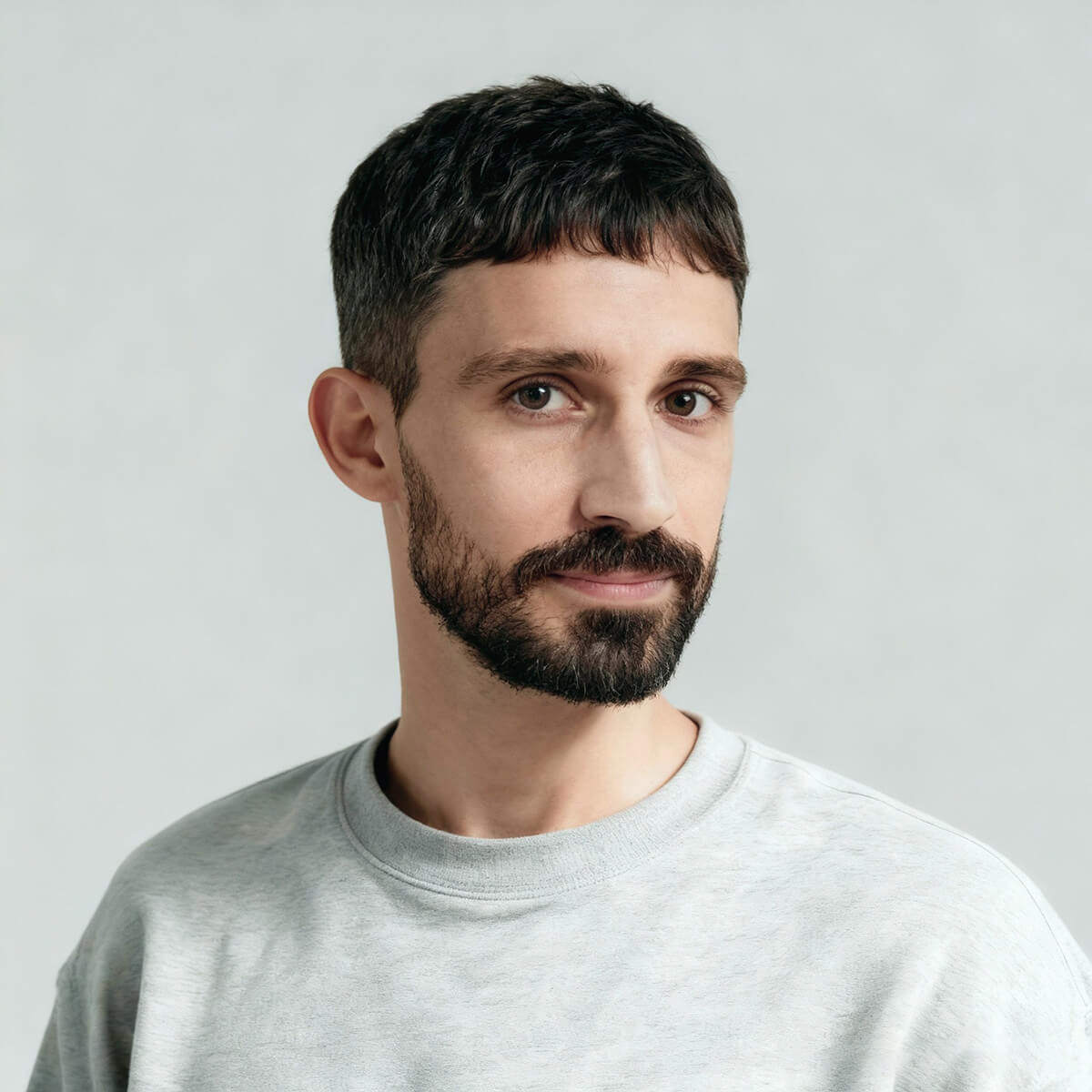 Bearded man with short dark hair wearing a light gray sweatshirt against a plain light background.
