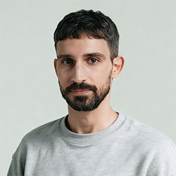 Man with short dark hair and beard wearing a light gray crewneck sweater against a plain light background.